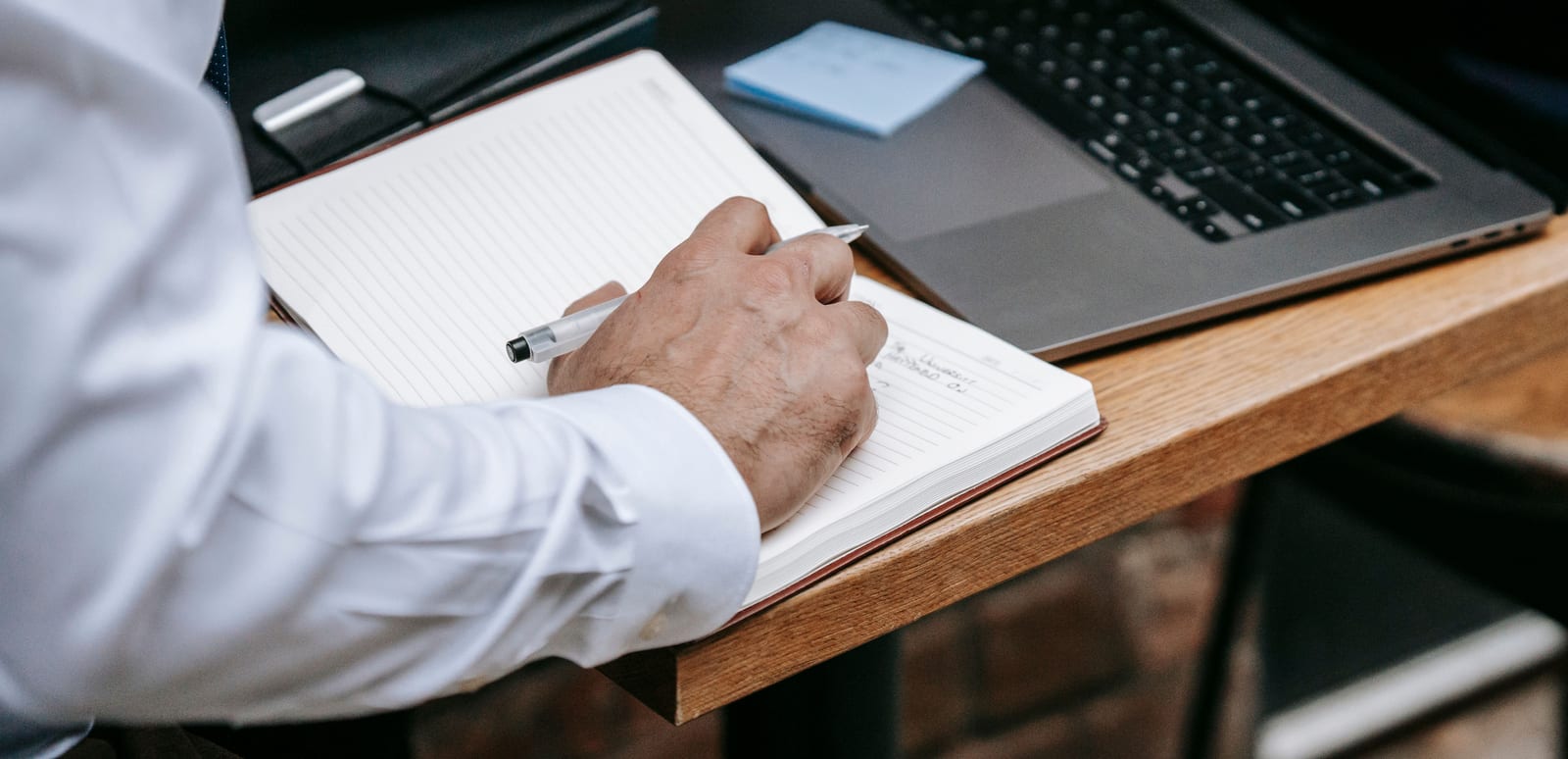 Person writing a list in a notebook next to an open laptop